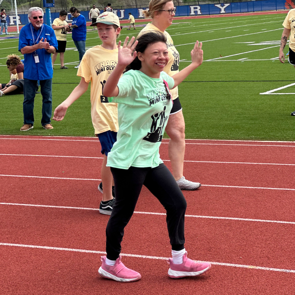 elementary student in light green t-shirt participating in the Wharton Mini Olympics outdoors on a track, smiling and running alongside a another student and a teacher.