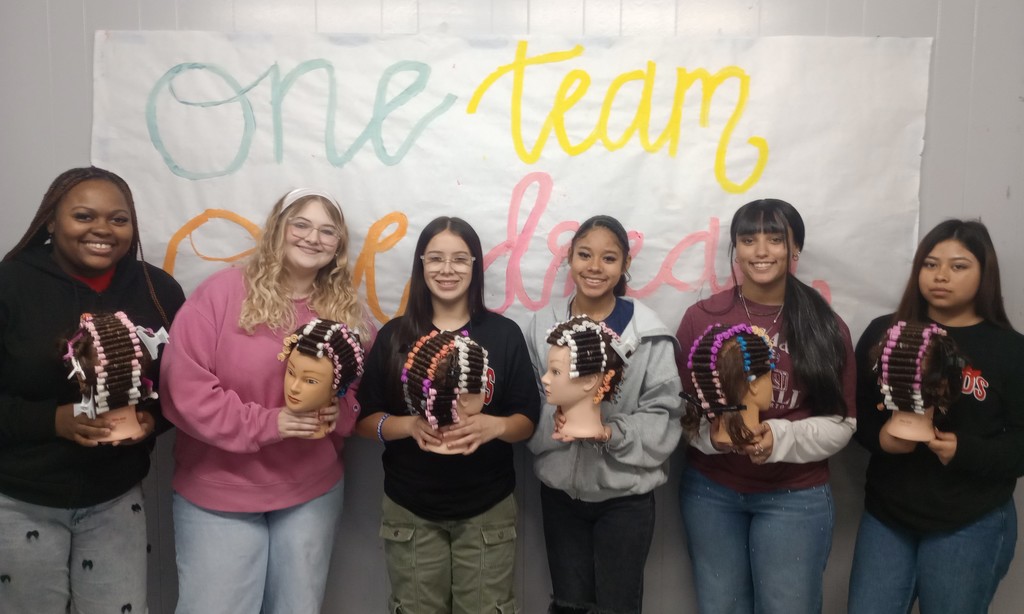 A group of Cosmetology 1 and 2 students at El Campo High School smiling and posing together while holding mannequin heads featuring their first completed full perm rod sets.