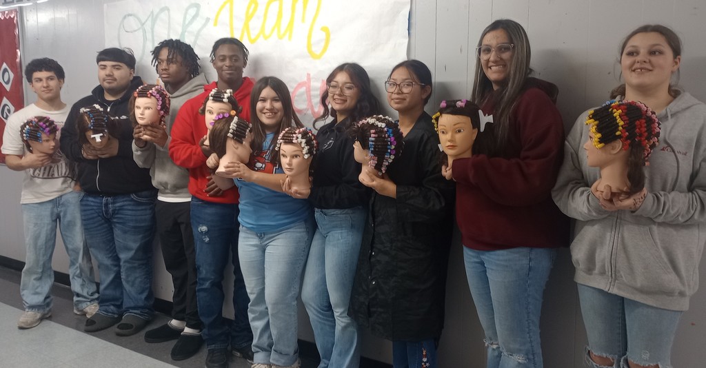 A group of Cosmetology 1 and 2 students at El Campo High School smiling and posing together while holding mannequin heads featuring their first completed full perm rod sets.