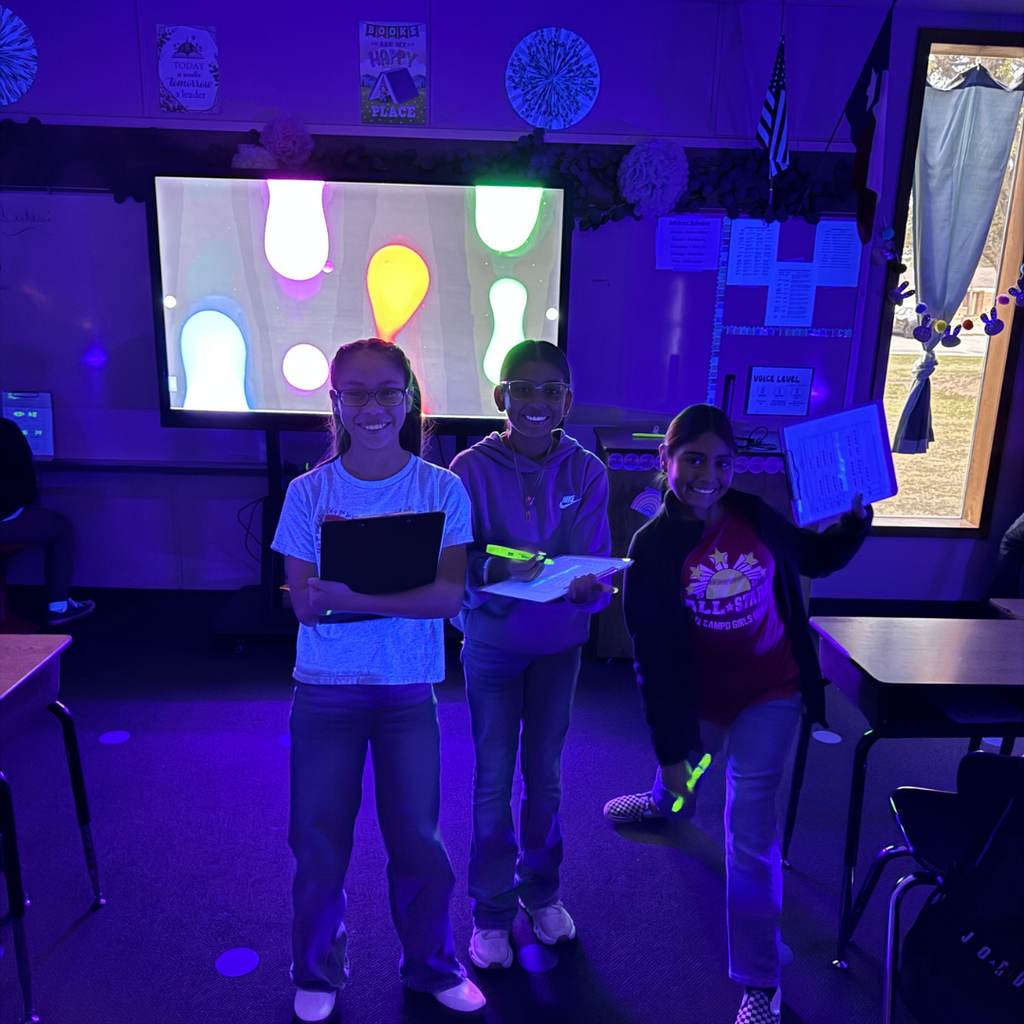 Three children stand in a darkened classroom illuminated by purple light. A screen behind them displays colorful, abstract shapes. 