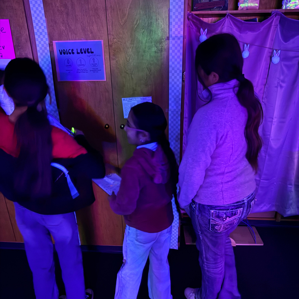 Three individuals stand near a wooden wall with a word problem posted. The room is illuminated with purple lights.