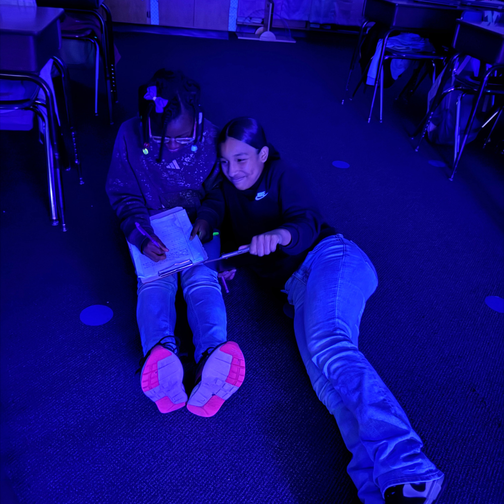 Two individuals are sitting on the floor in a room illuminated by blue light, with one holding a clipboard and the other holding a pencil .