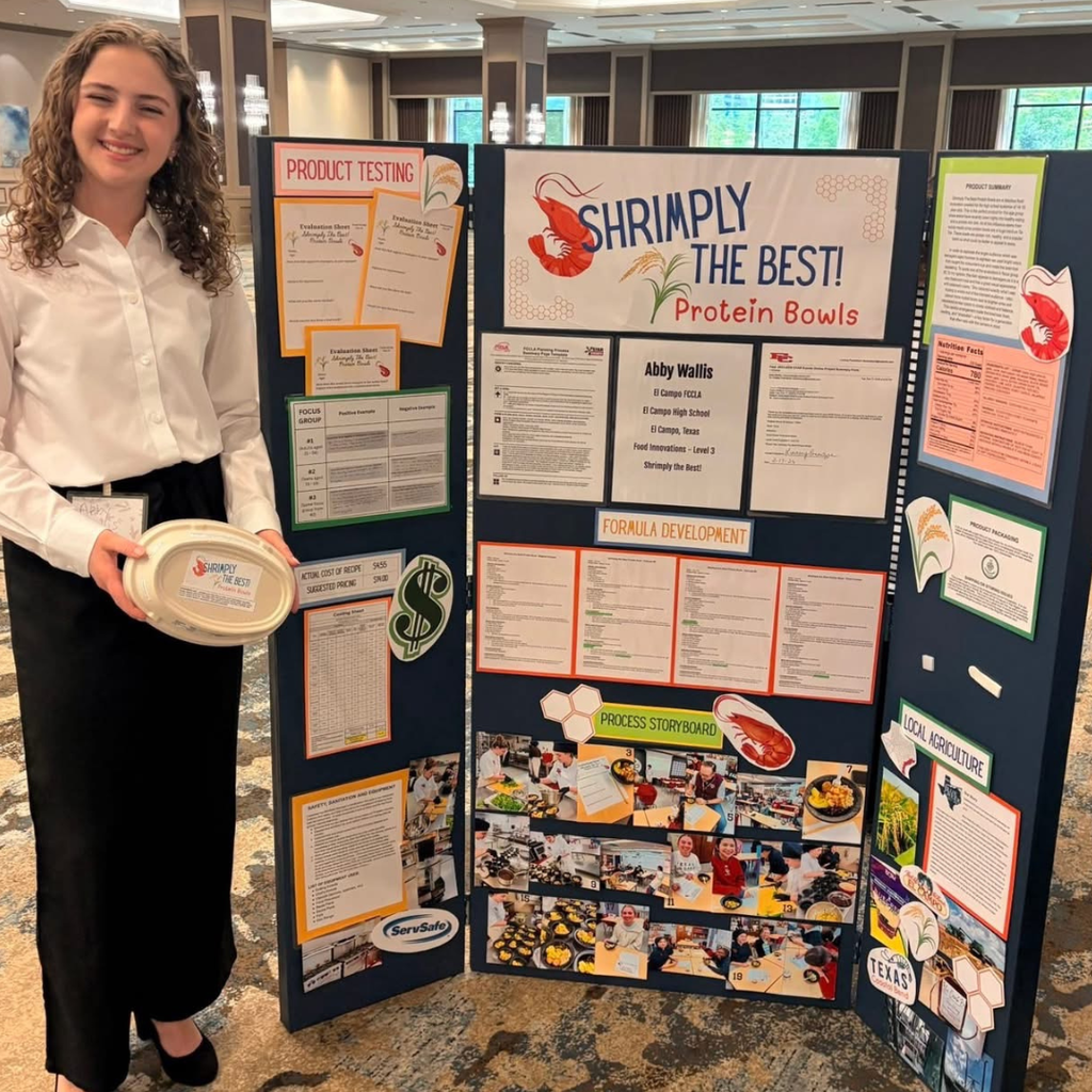 Wallis stands next to a board titled "SHRIMPLY THE BEST! Protein Bowls" which displays information about product testing, research, and development. Wallis is holding a white oval container with a "shrimply the best label