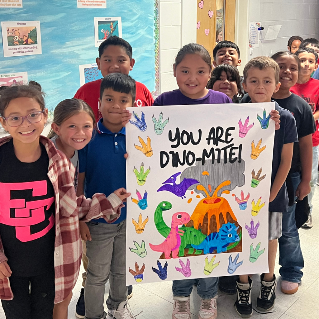 A classroom photo of students smiling behind a "You are DINO-MITE!" encouragement sign decorated with dinosaurs and a volcano.