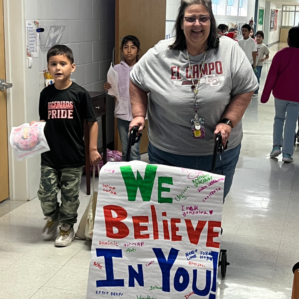 A teacher and student holding a colorful "We Believe In You!" poster covered in student names in a school hallway.