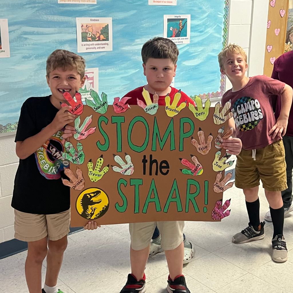 Three elementary students holding a large poster that says "STOMP the STAAR!" in bold green letters. The poster is decorated with a T-Rex footprint and numerous colorful paper handprints, each featuring a student's name