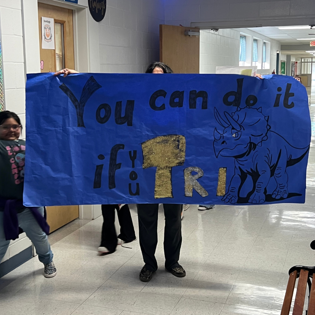 Two students in a school hallway holding a large dark blue poster that reads "You can do it if you TRI!" The word "TRI" is highlighted in yellow and decorated with a drawing of a blue triceratops.