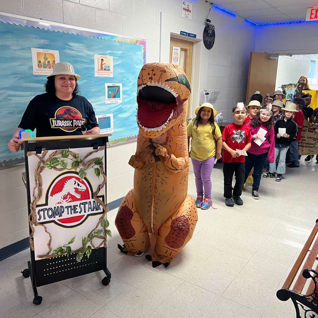 A staff member in a Jurassic Park shirt and hat stands next to a podium that says "Stomp the STAAR." Beside her is a person in an inflatable T-Rex costume, with a line of elementary students waiting in the school hallway.