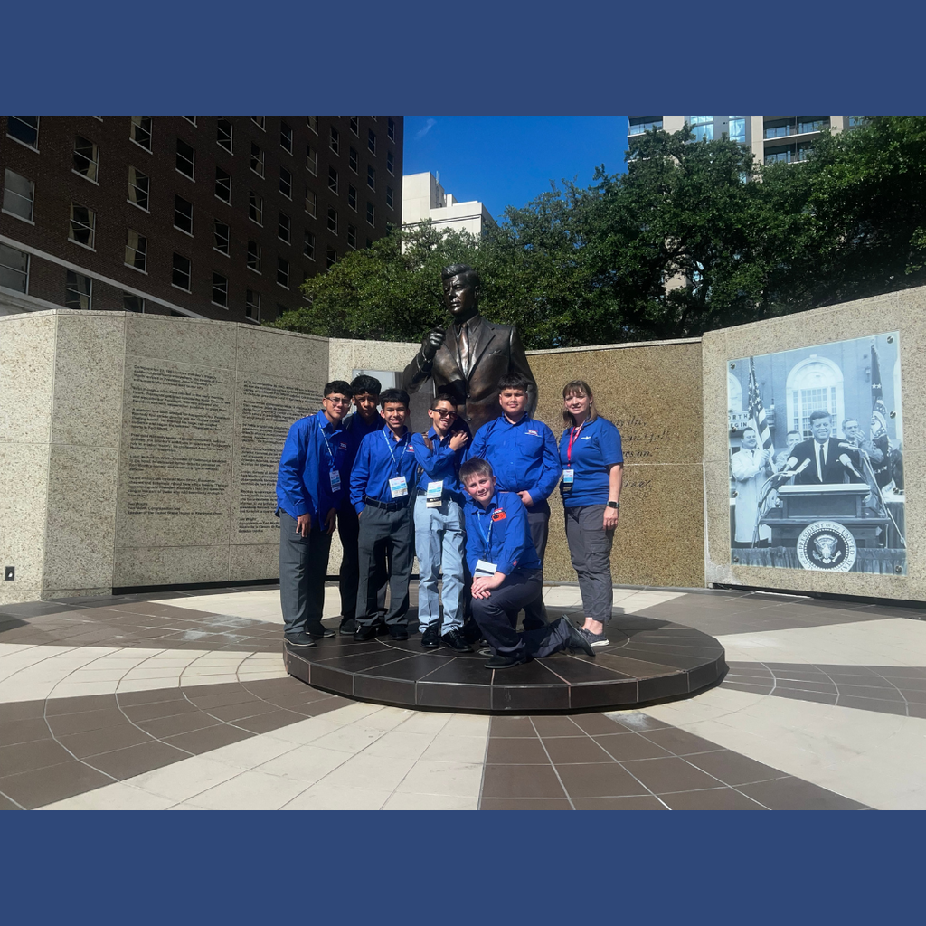 6 kids and TSA sponsor in blue shirts pose for a photo in front of a statue of John F. Kennedy in Dallas.