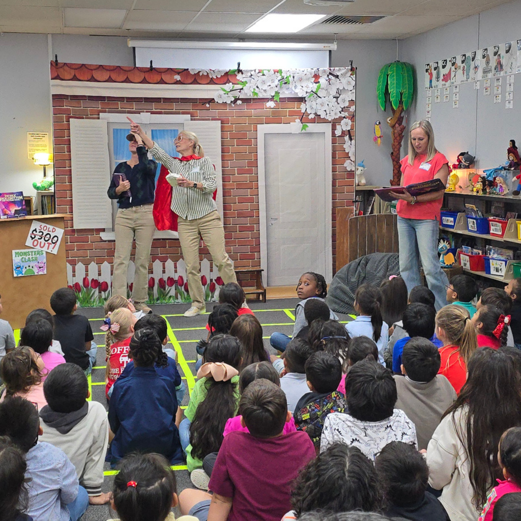 Members of the Sesame Club wearing red capes engage with Myatt students during a Reading Is Fundamental (RIF) book party in a decorated library setting.