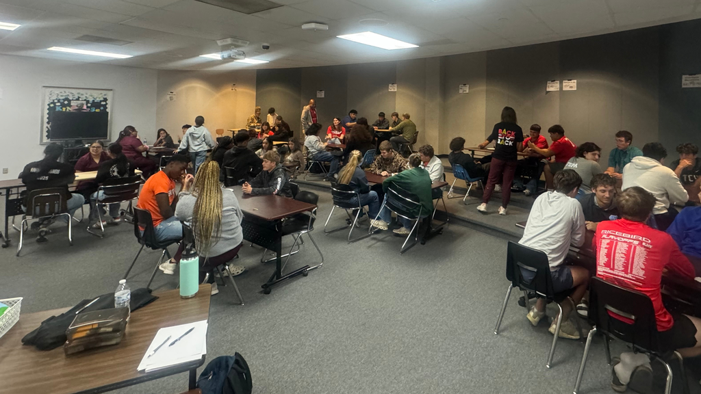 A large classroom filled with high school students sitting at clustered desks, working together in small groups and focusing on their materials during a history review session.