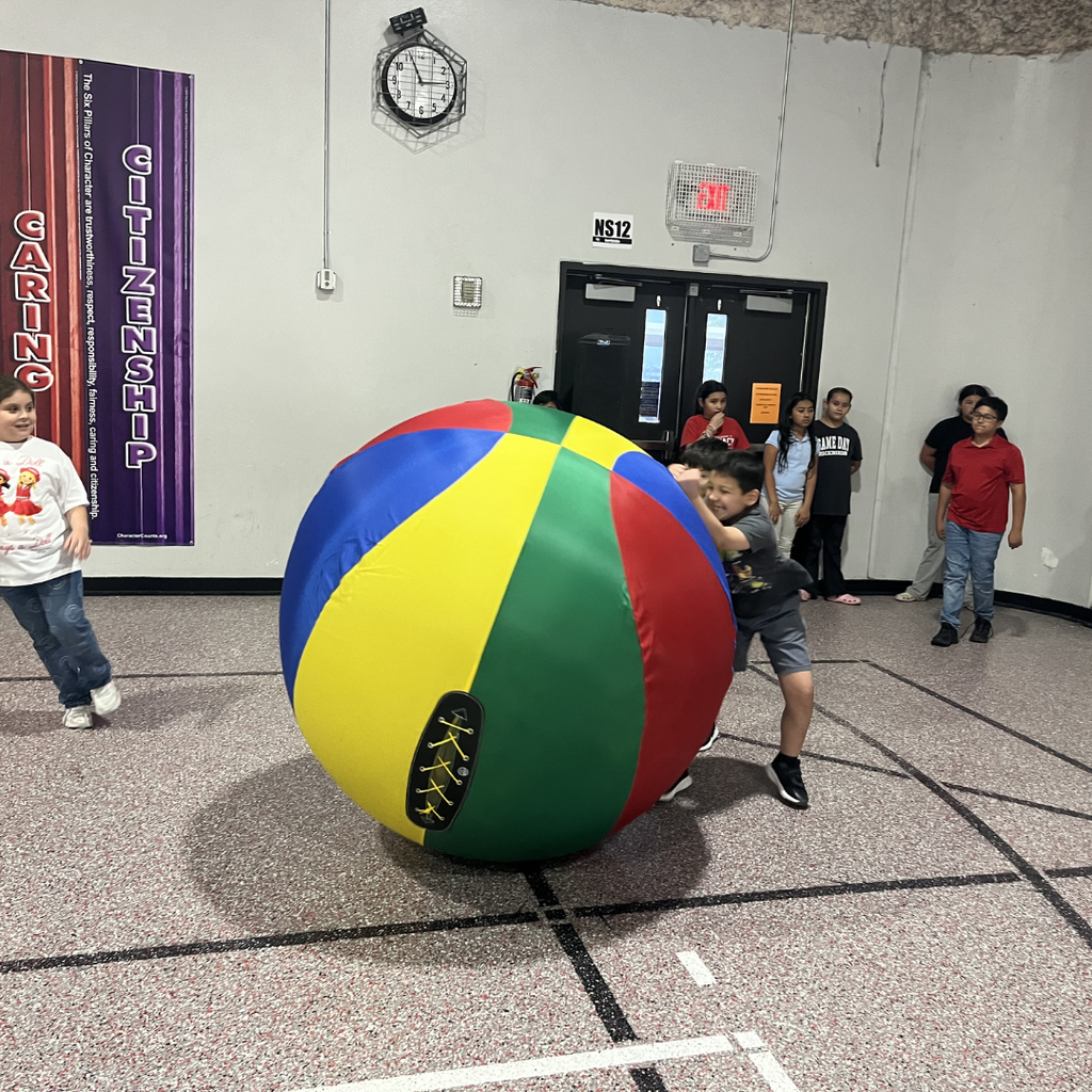 A student in a gymnasium preparing to roll a large, blue inflatable ball toward oversized white bowling pins