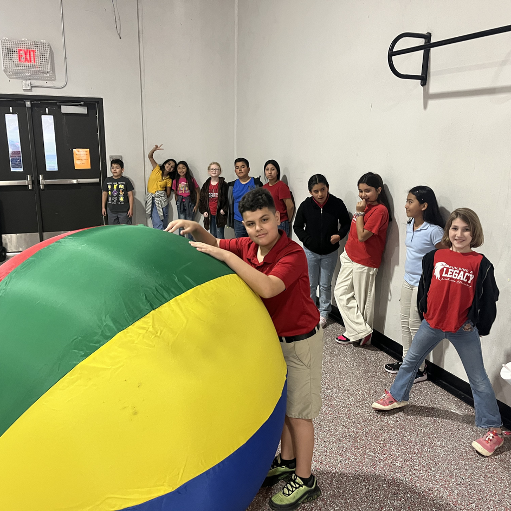 A student in a gymnasium preparing to roll a large, blue inflatable ball toward oversized white bowling pins