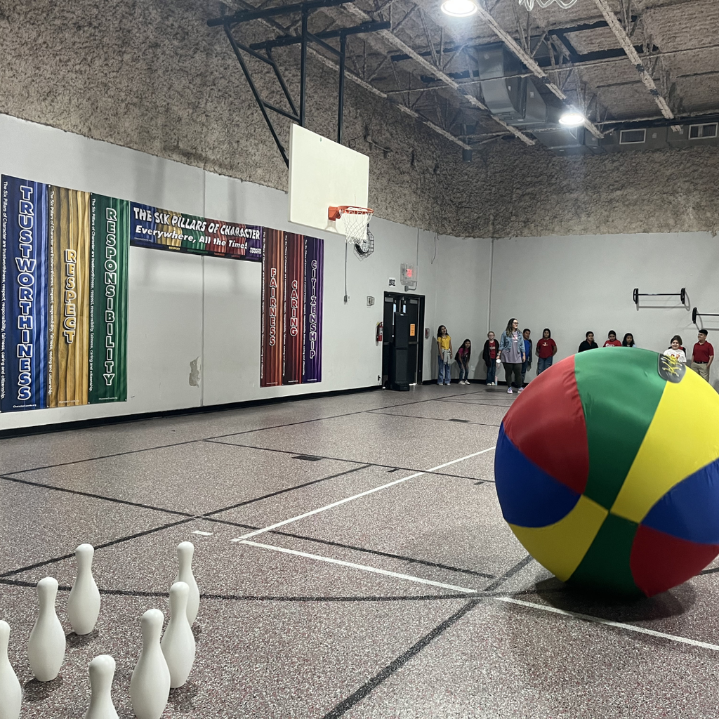 A student in a gymnasium preparing to roll a large, blue inflatable ball toward oversized white bowling pins