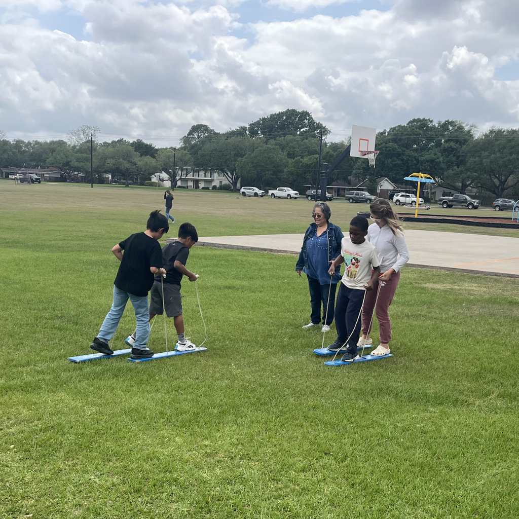 Two pairs of students practicing coordination on blue team-walking planks outdoors.