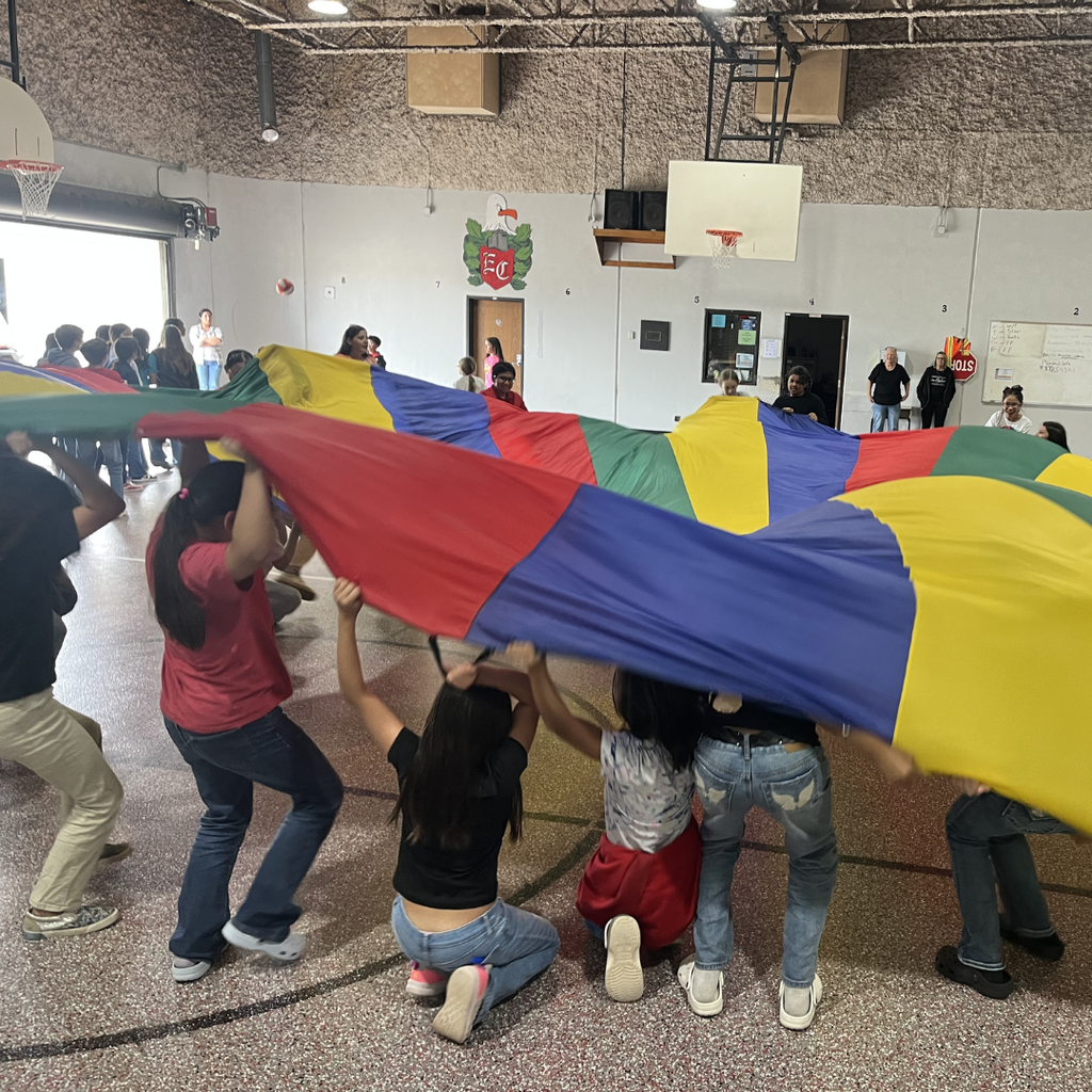 A group of students in a gymnasium lifting a large, colorful multicolored parachute high into the air