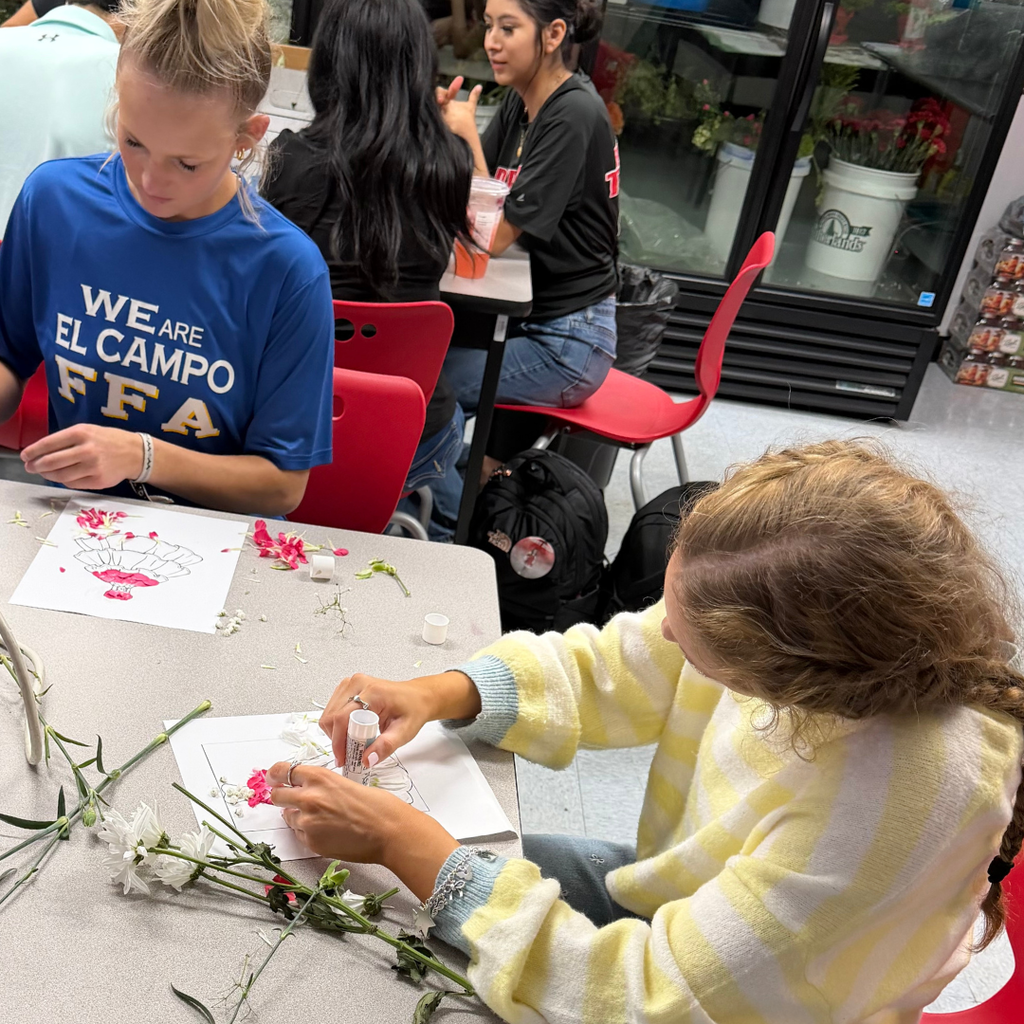 Two Advanced Floral Design students sitting at a table, carefully glueing real flower petals onto a paper dress template to create a floral fashion design.