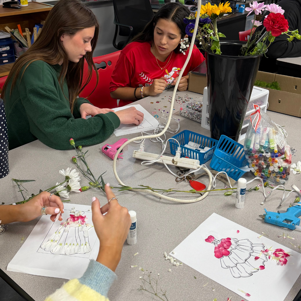Two Advanced Floral Design students sitting at a table, carefully glueing real flower petals onto a paper dress template to create a floral fashion design.