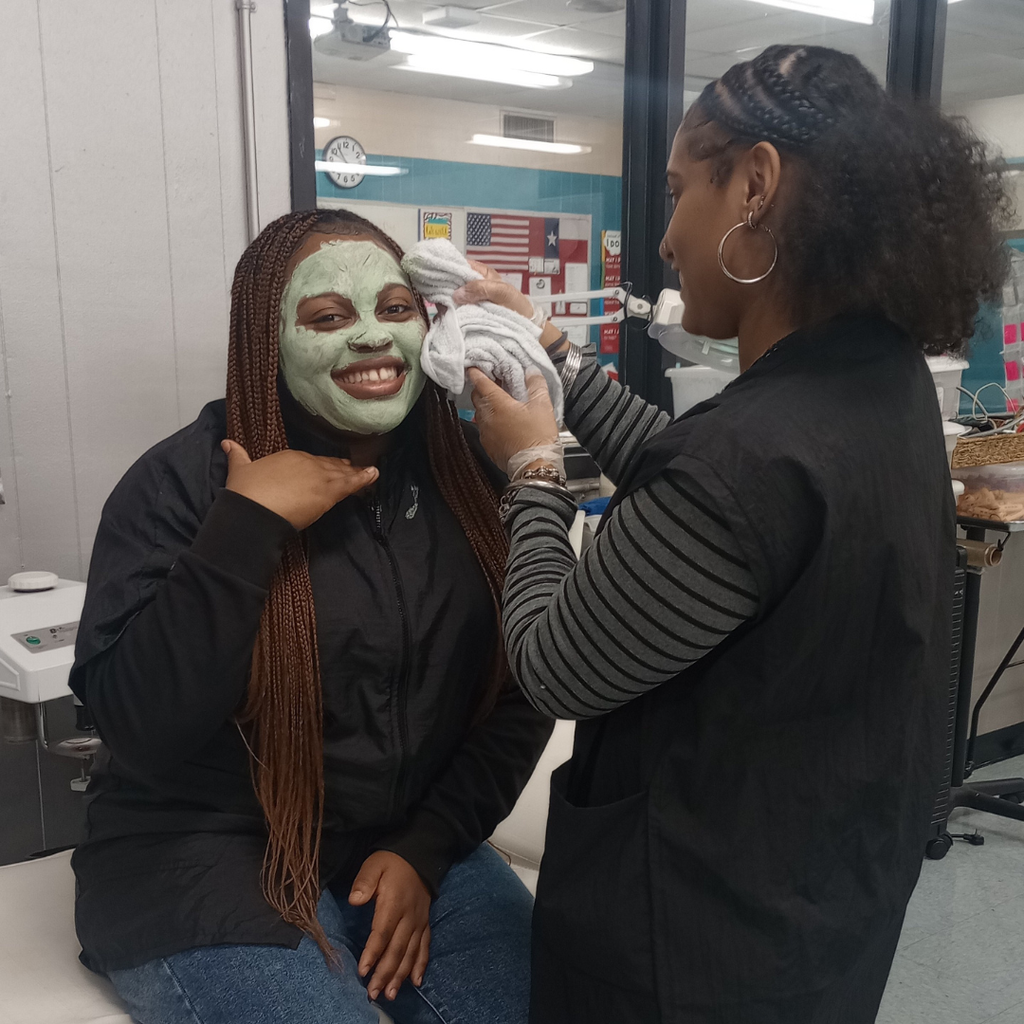 student with a green facial mask smiles at another student begins to wipe the mask off with a towel