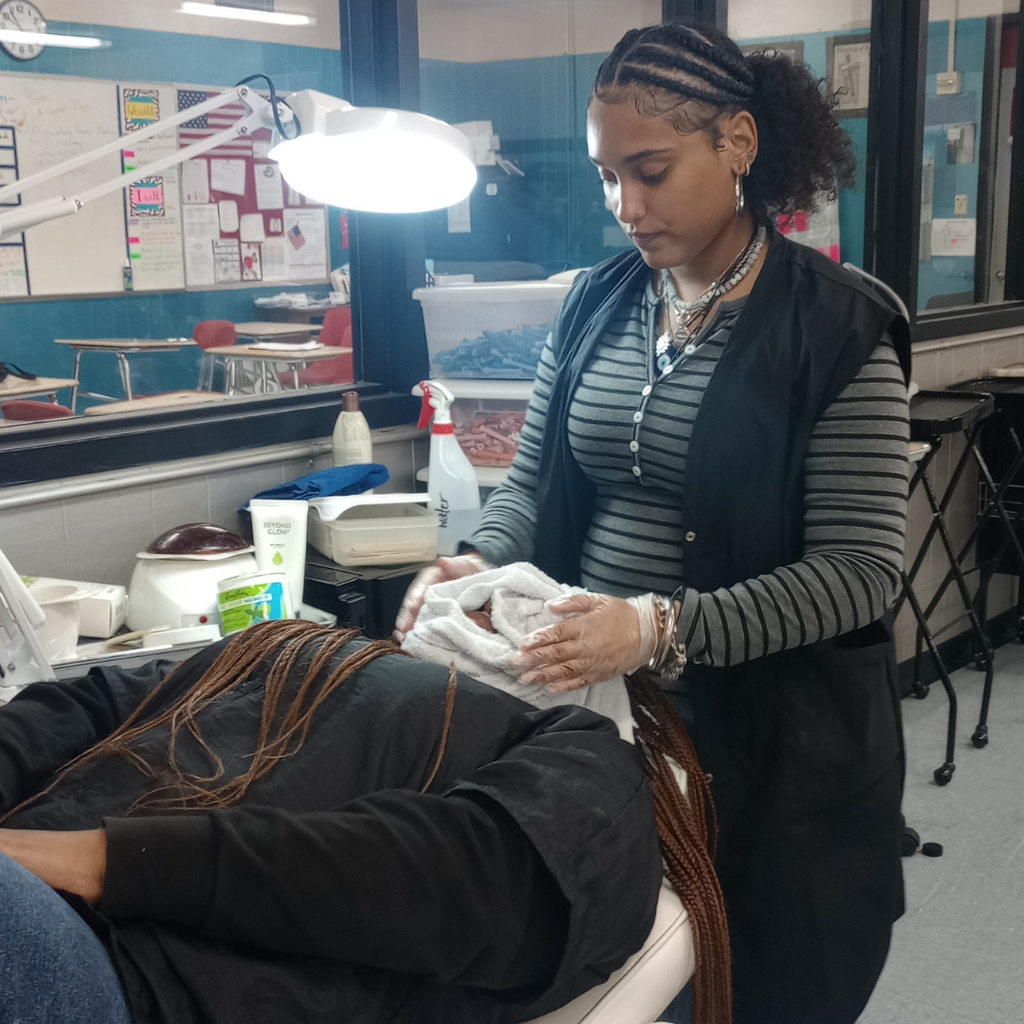 A cosmetology student, Paige Means, wearing a striped shirt and black vest, carefully applies a warm towel facial to another student, Ma'Leah Scarlett, who is reclining in a salon chair.