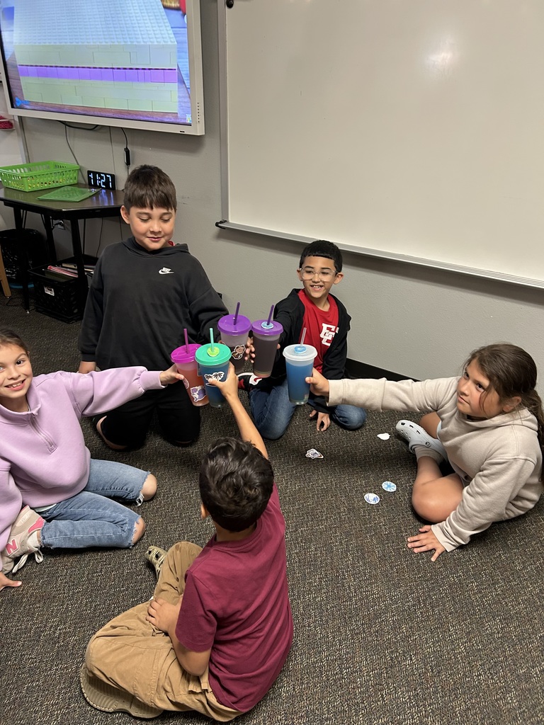 A group of elementary students sitting in a circle in a classroom, smiling and cheering as they "toast" by clinking their colorful reusable cups together in celebration.