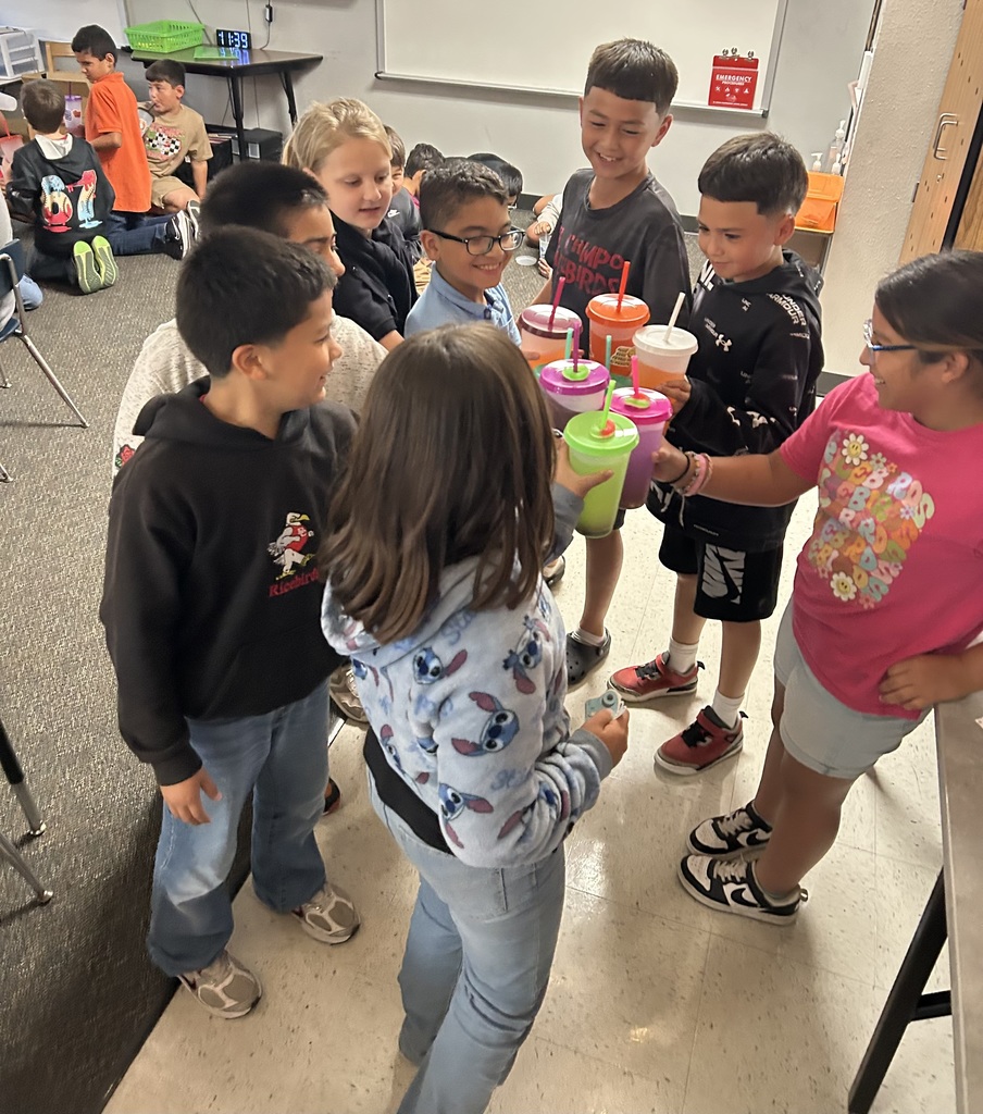 A group of elementary students standing in a circle in a classroom, smiling and cheering as they "toast" by clinking their colorful reusable cups together in celebration.