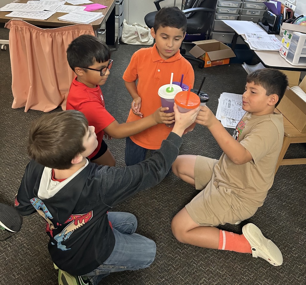 A group of elementary students sitting in a circle in a classroom, smiling and cheering as they "toast" by clinking their colorful reusable cups together in celebration.