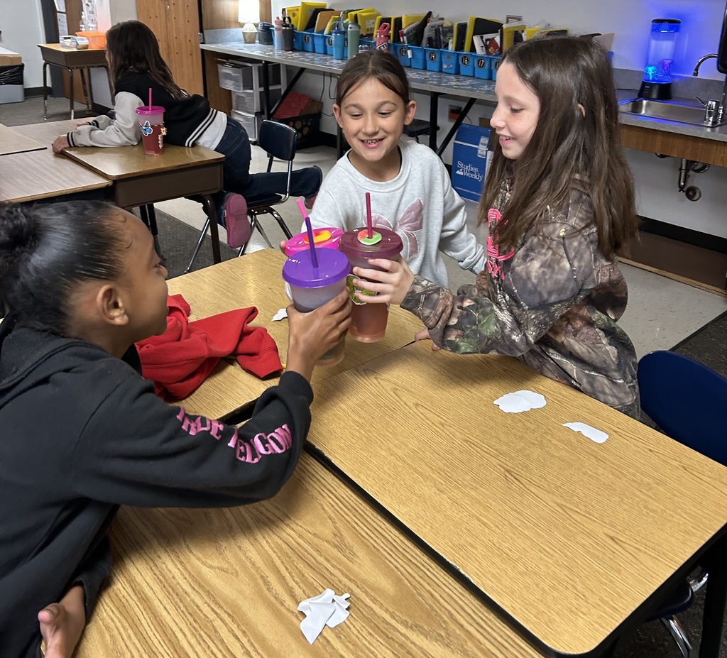A group of elementary students sitting at desks  in a classroom, smiling and cheering as they "toast" by clinking their colorful reusable cups together in celebration.