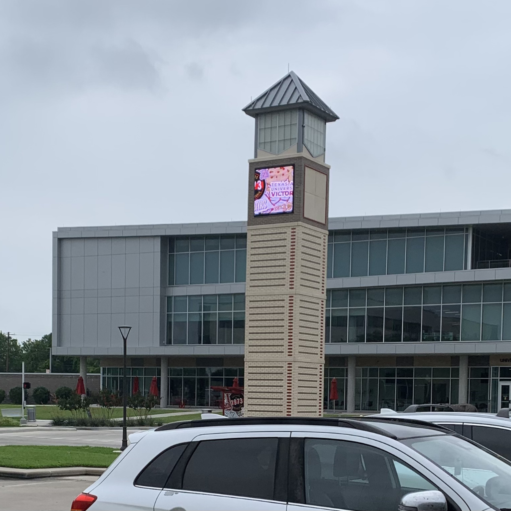 A tall brick clock tower featuring a digital sign for the University of Houston-Victoria, situated in front of a modern campus building under a cloudy sky.