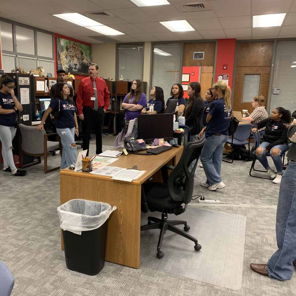 group of ECHS HOSA students standing in the counselor's office at the A&M campus, listening to a man in a red shirt standing near a desk in the center, and several students in blue HOSA shirts are gathered around.