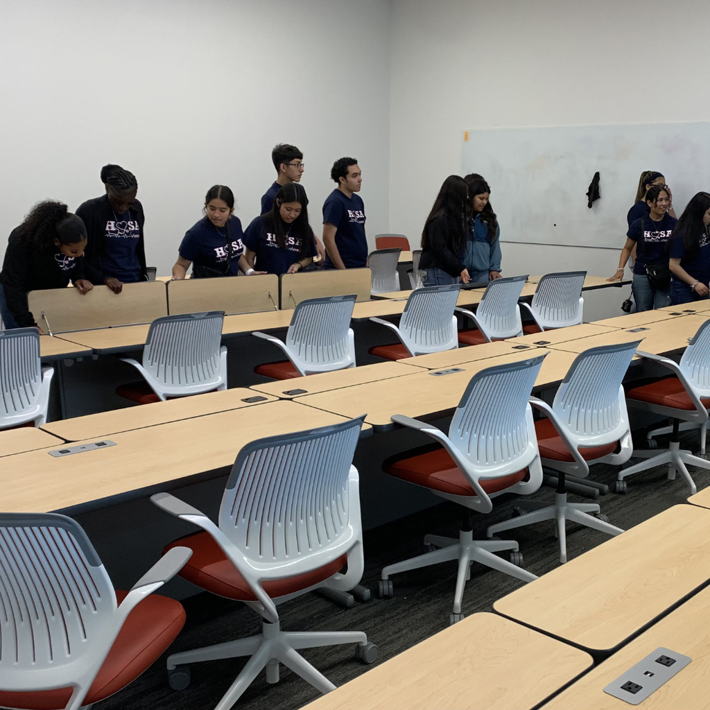 A group of ECHS HOSA students standing and talking in a modern university classroom with rows of white chairs and wooden tables.