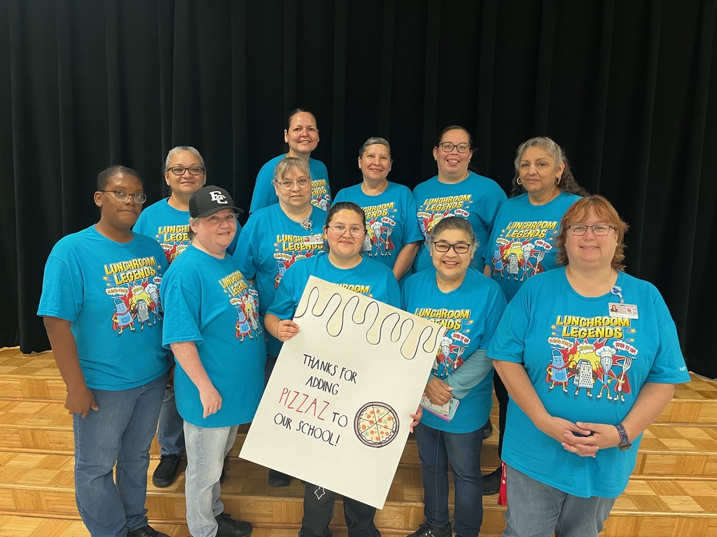 A group of ten school cafeteria staff members wearing matching bright blue "Lunchroom Legends" t-shirts. They are standing together on a stage in front of a black curtain, smiling and holding a handmade sign that reads, "Thanks for adding PIZZAZ to our school!"