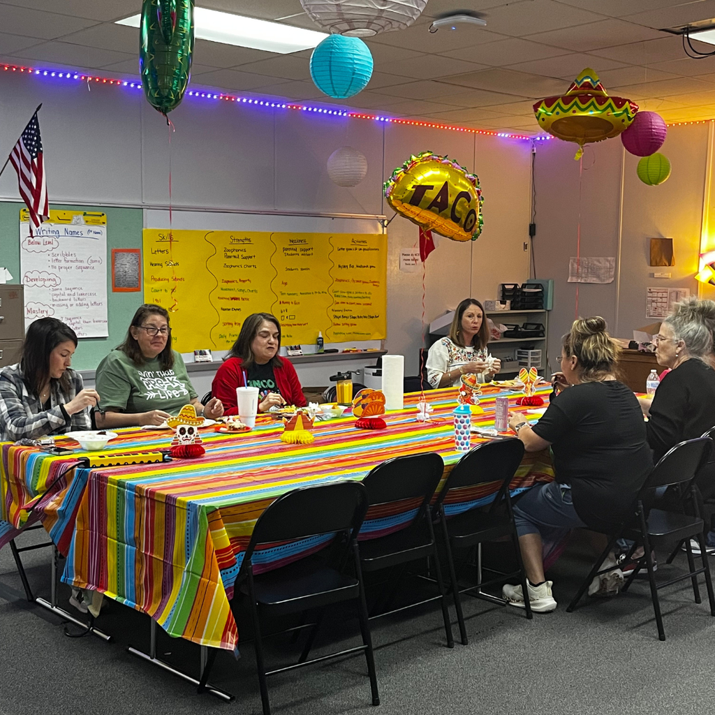 A group of school staff members sitting around a long table decorated with a colorful striped Mexican serape tablecloth. The room is decorated with festive paper lanterns and a "TACO" balloon for a luncheon.