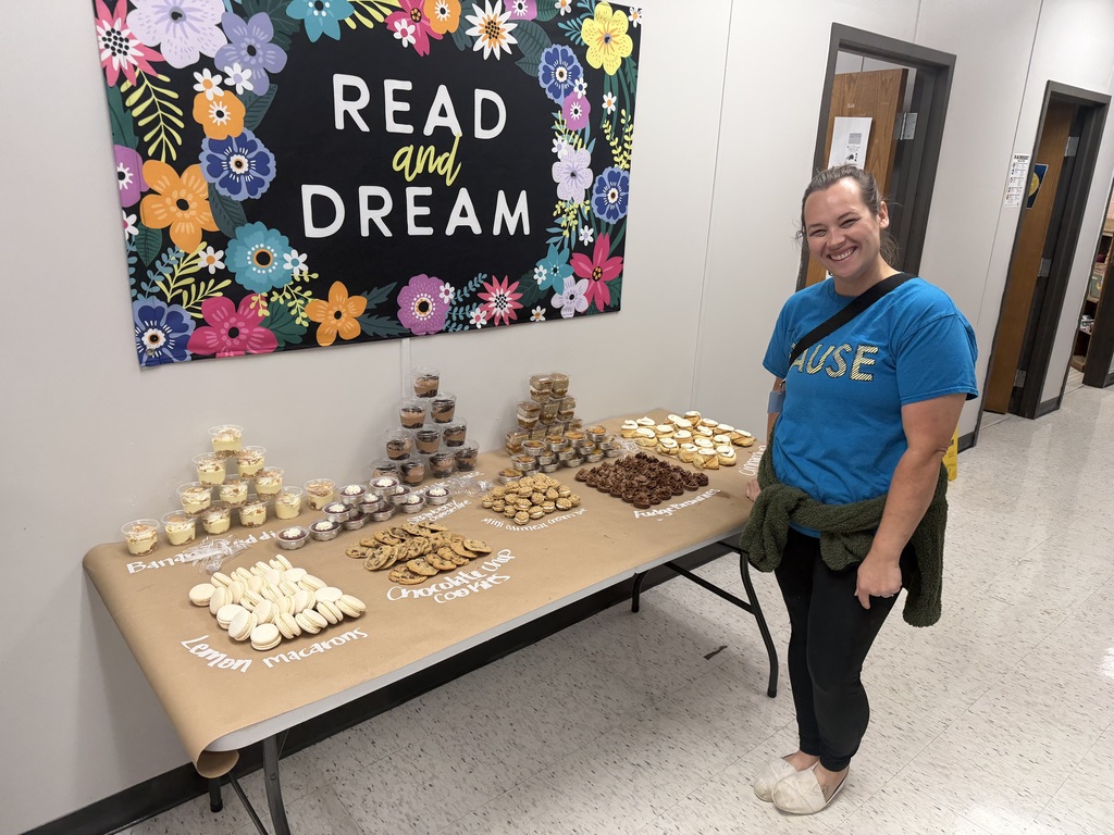 A staff member stands next to a dessert table at Northside Elementary featuring a "Read and Dream" floral banner. The table is filled with organized trays of macarons, chocolate cookies, and cupcakes.