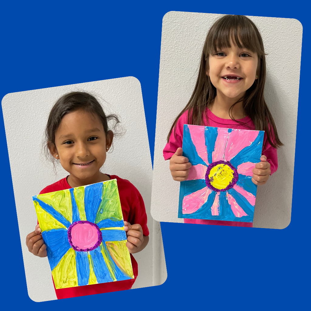 kindergarten students stand together smiling and holding up their colorful, hand-painted flower artwork. The students are each proudly displaying a unique bluebonnet-inspired painting featuring vibrant petals and bright backgrounds.