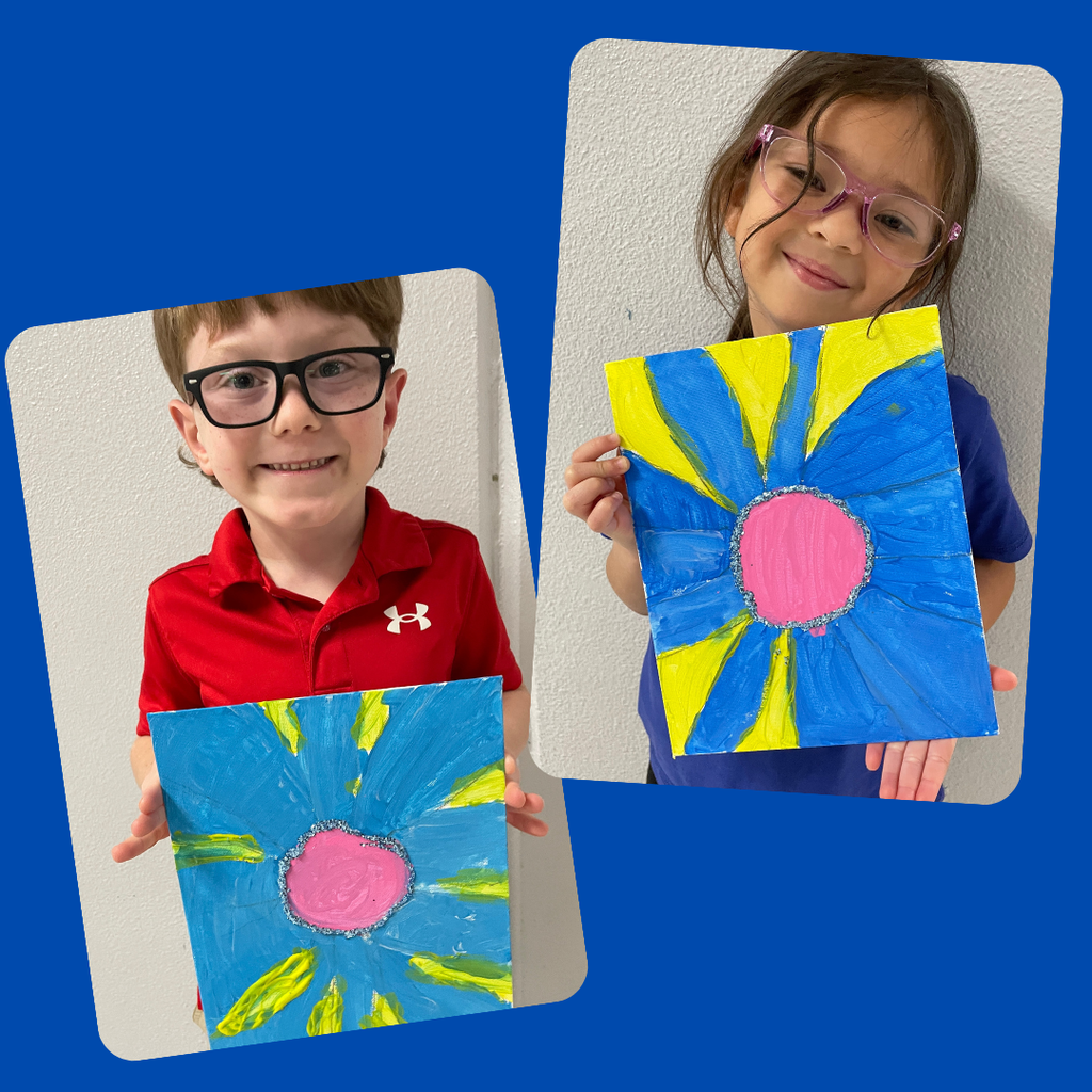 kindergarten students stand together smiling and holding up their colorful, hand-painted flower artwork. The students are each proudly displaying a unique bluebonnet-inspired painting featuring vibrant petals and bright backgrounds.