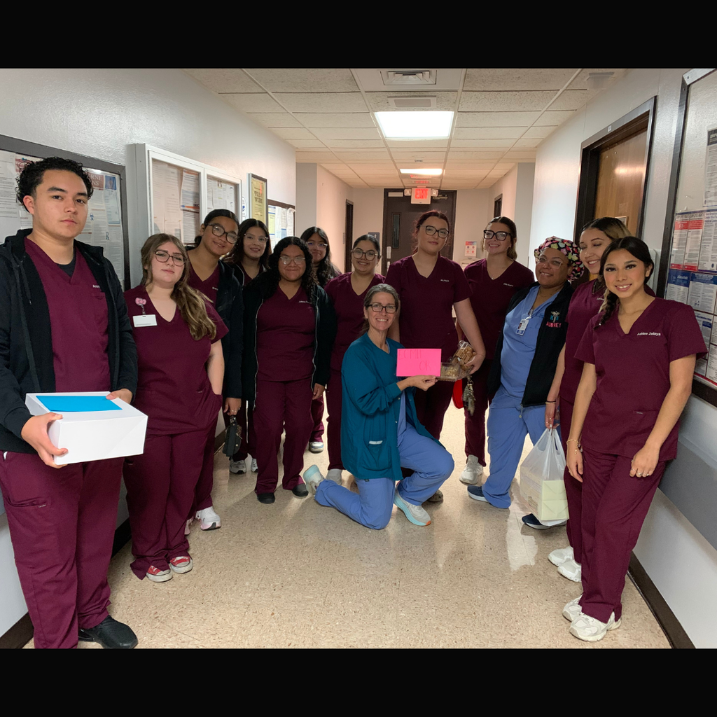 students in maroon scrubs standing in a clinical office setting, presenting a handmade thank-you card and a tray of baked goods to a healthcare professional.