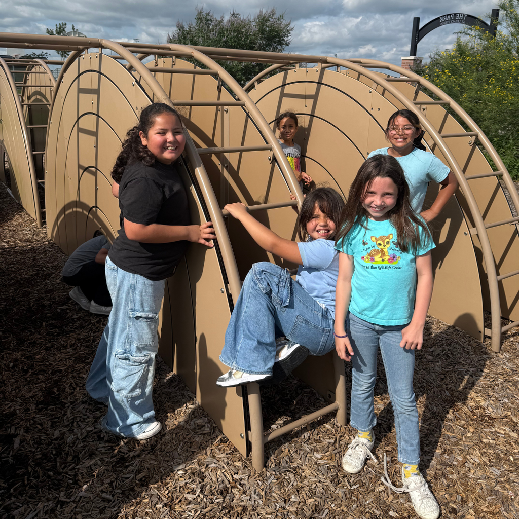 Three smiling 3rd-grade girls posing together while climbing on a tan, curved playground structure at the park