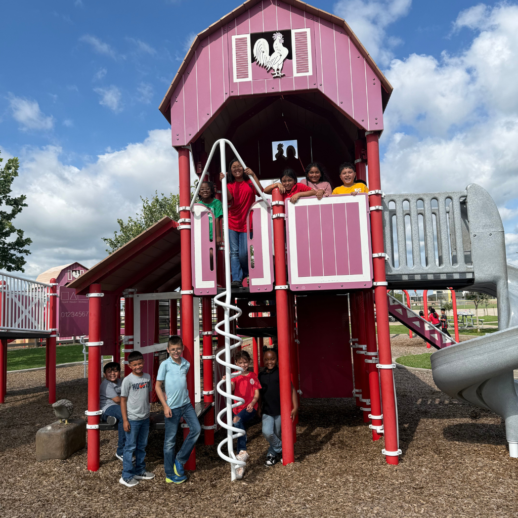 A group of elementary students posing on a large red and white barn-themed playground structure. Some students are standing on the ground in front, while others are waving from the upper deck of the playhouse under a blue sky with white clouds