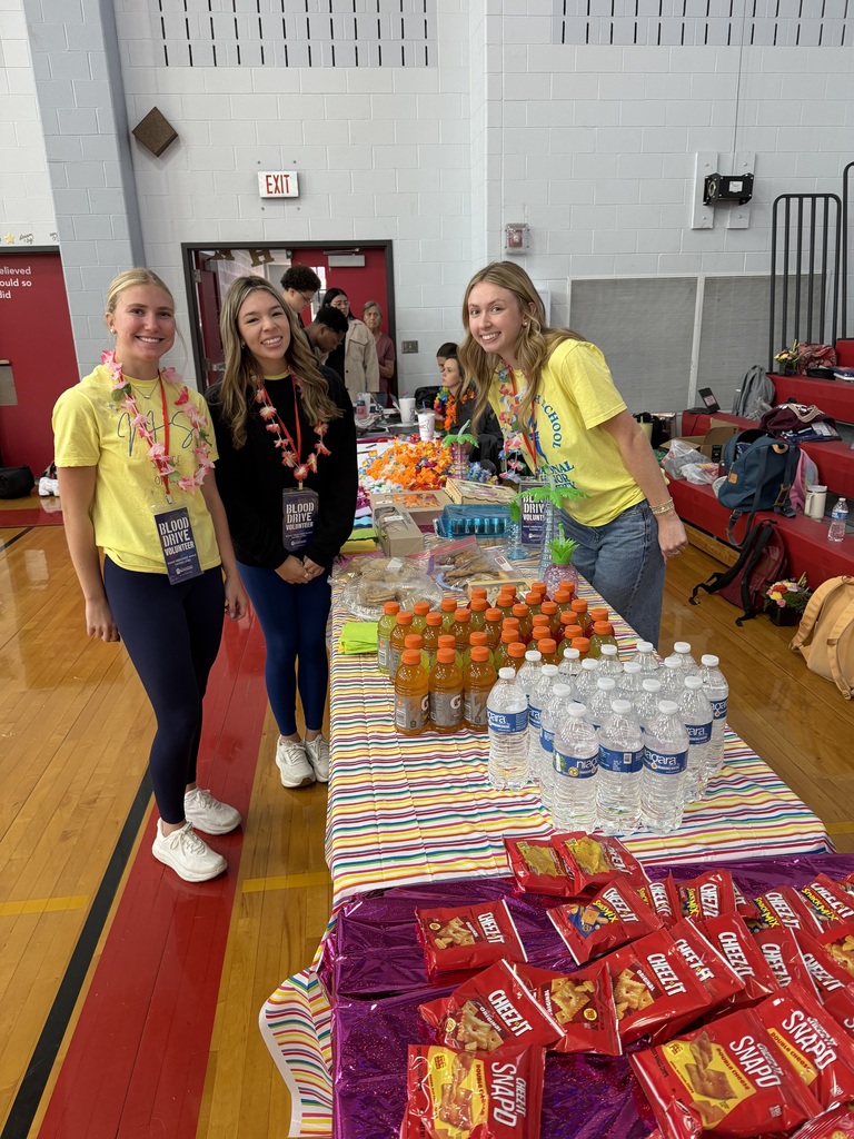 3 students at the blood drive wearing leis standing by food and drink table