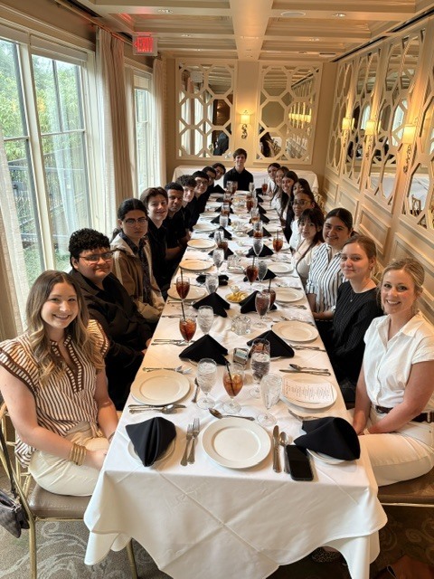A group of high school culinary arts students smiling and posing together around a long dining table inside a beautifully decorated, sunlit restaurant.