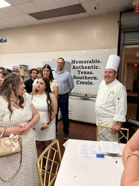 In a bright commercial kitchen, a chef in a white coat stands on the left, gesturing with an raised hand as he addresses a line of high school culinary students listening attentively