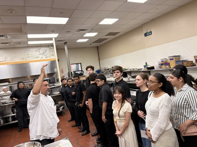 In a bright commercial kitchen, a chef in a white coat stands on the left, gesturing with an raised hand as he addresses a line of high school culinary students listening attentively