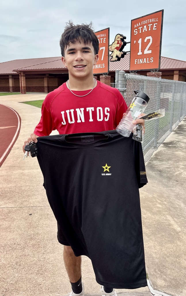 A young male Ricebird soccer player stands on an outdoor track in front of a school building. He is wearing a red shirt that says "JUNTOS" and holding up a black U.S. Army t-shirt featuring a gold star. He is smiling and holding a water bottle in his other hand.