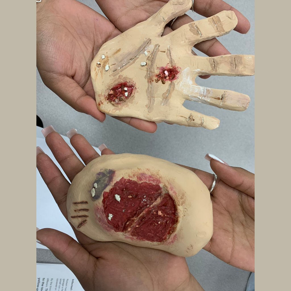 Hands holding two clay models created by health science students to simulate trauma wounds, including a clay hand with lacerations and an oval clay model showing a deep open wound with exposed red tissue.
