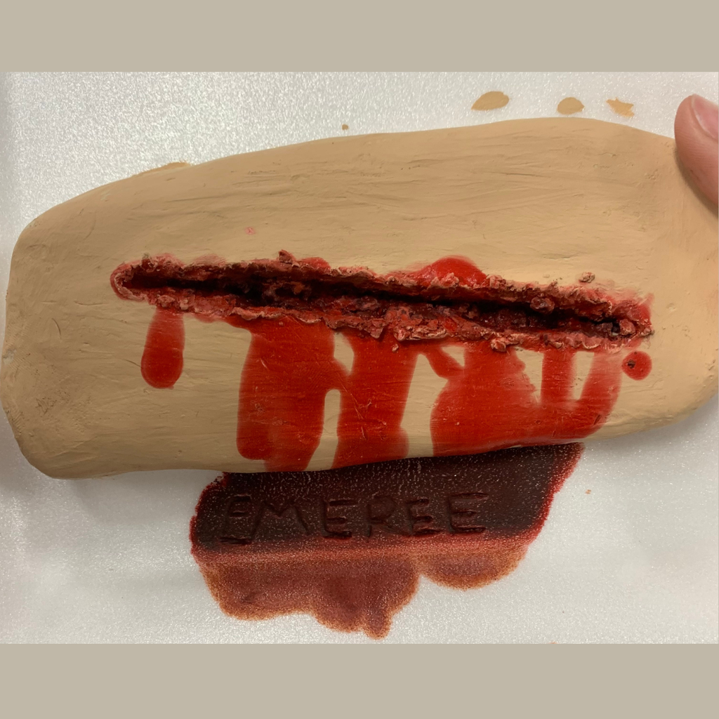 A close-up of a hands-on classroom project showing a flat piece of beige clay sculpted to look like a deep skin wound. The center features a long, textured laceration filled with realistic dark red faux blood, with multiple streaks of blood dripping vertically down the clay surface.