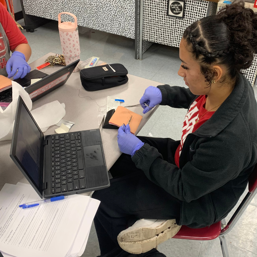 A student wearing purple gloves uses surgical tools to practice stitching a suture pad during a health science class.