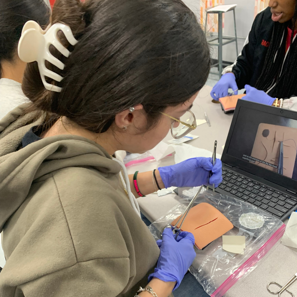 A student wearing purple gloves uses surgical tools to practice stitching a suture pad during a health science class.