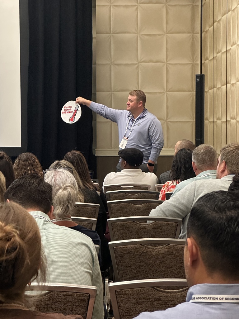 An El Campo Middle School administrator stands at the front of a presentation room at the TASSP conference, holding up a round prop to emphasize a point to an audience seated in rows.