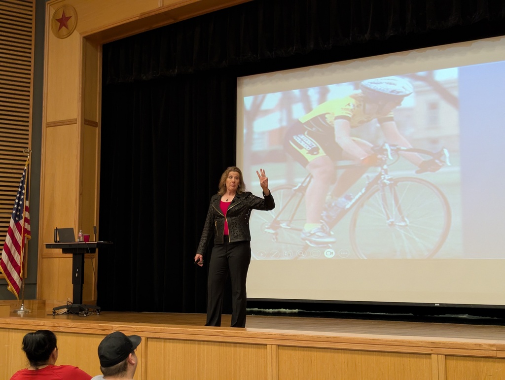 brown on stage talking with a picture of her on a bike in the background